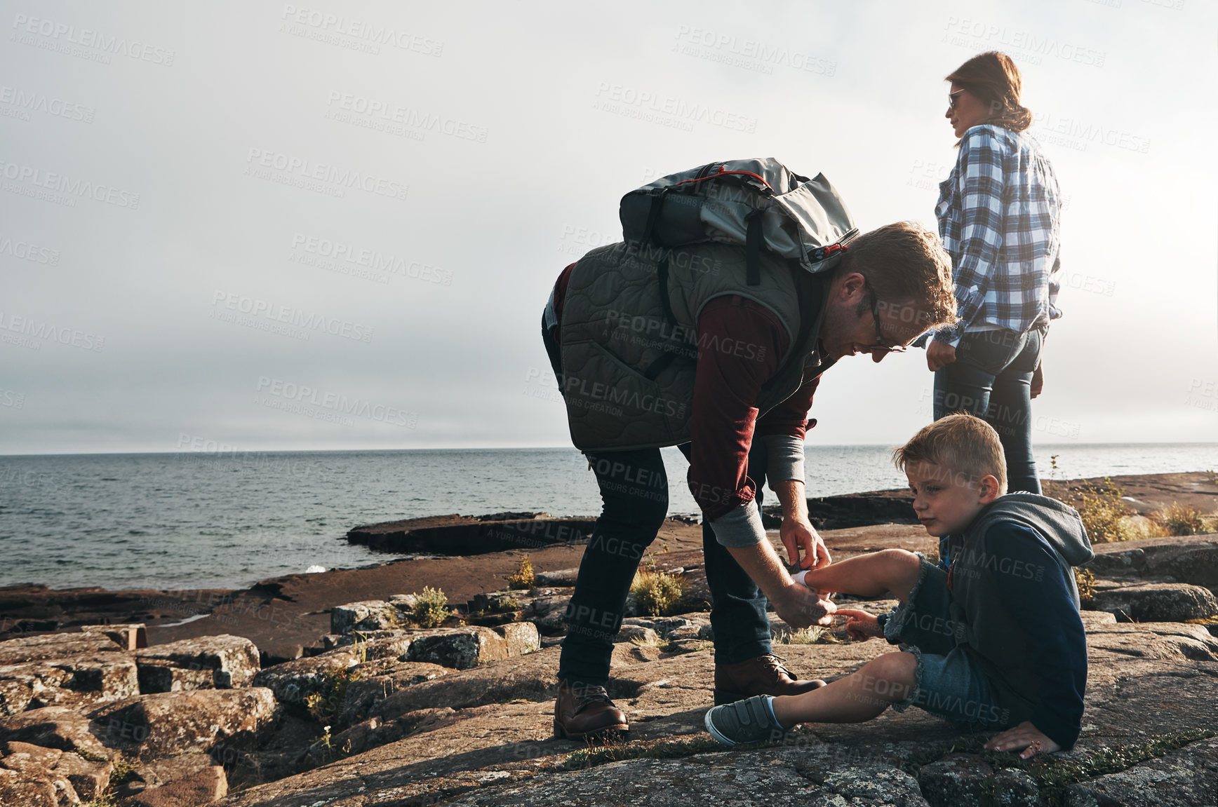 Buy stock photo Shot of a cheerful man helping to put his son's shoes on while standing next to the ocean outside during the day