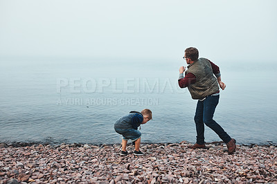 Buy stock photo Rearview shot of a cheerful father and son throwing flat stones into a lake while standing next to the water