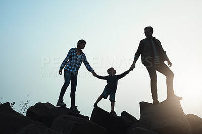 Buy stock photo Wide shot of a silhouette of a young family holding hands while walking on rocks with the sun peeking out in the horizon