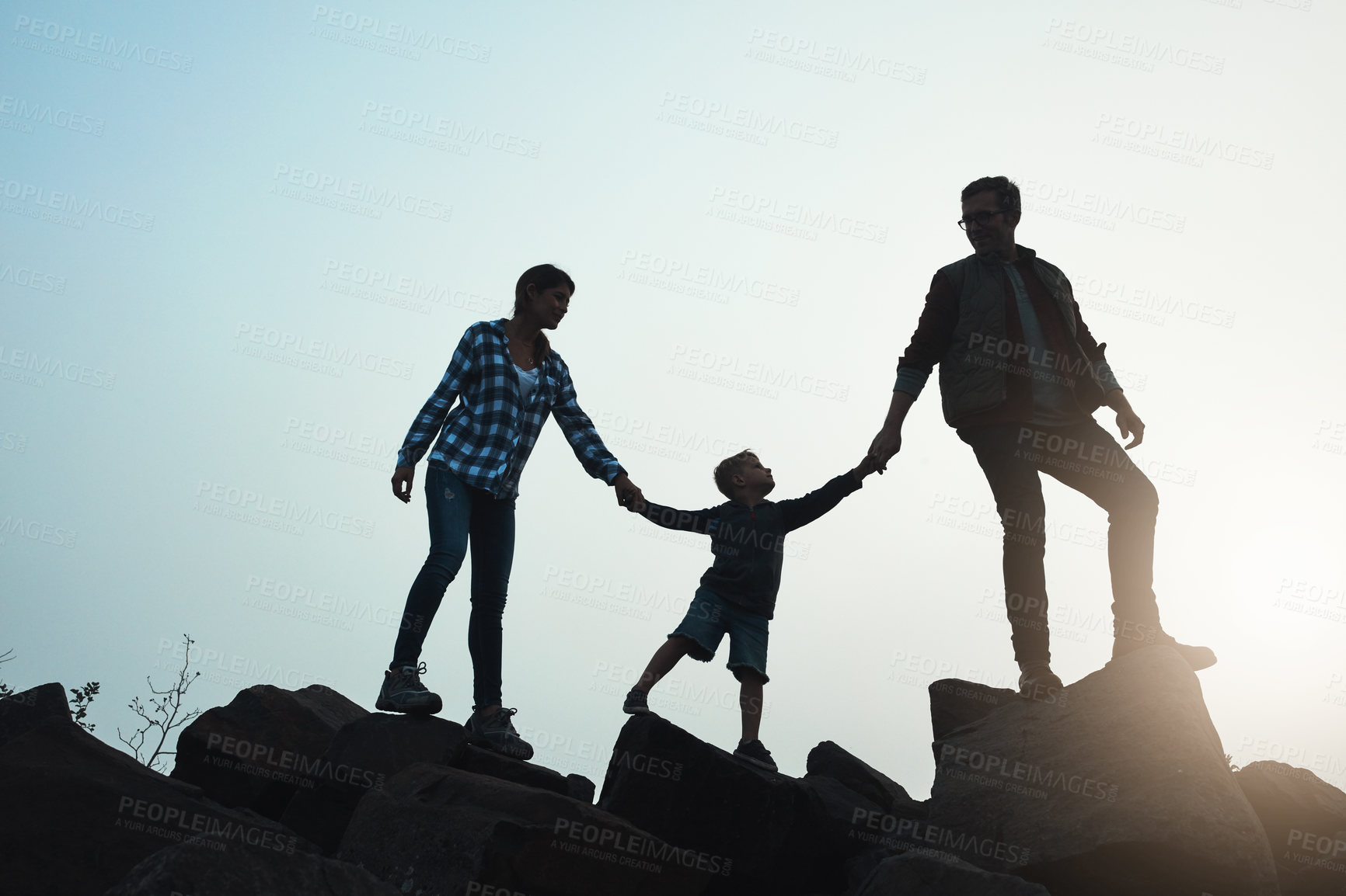 Buy stock photo Wide shot of a silhouette of a young family holding hands while walking on rocks with the sun peeking out in the horizon