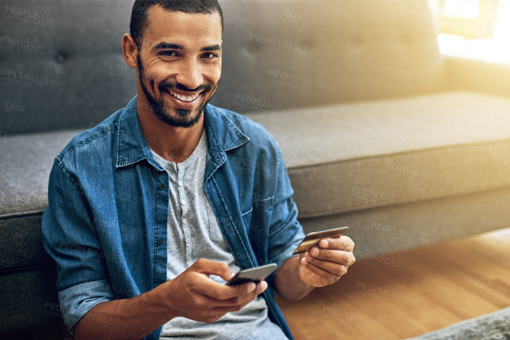 Buy stock photo Shot of a focused young man doing online banking with her phone while being seated on the floor next to a couch at home