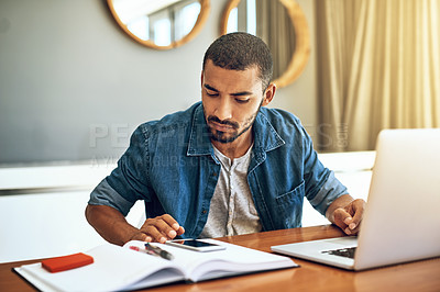 Buy stock photo Shot of a focused young man working on his laptop and checking his phone while being seated at a table at home