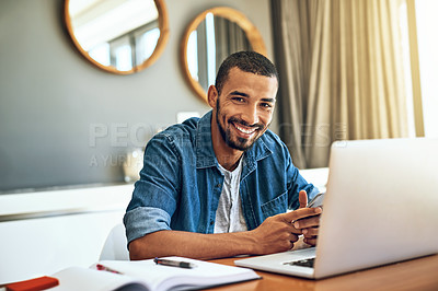 Buy stock photo Portrait of a cheerful young man working on his laptop and checking his phone while being seated at a table at home