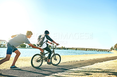 Buy stock photo Cropped shot of a young boy teaching his younger brother how to ride a bike alongside a lagoon