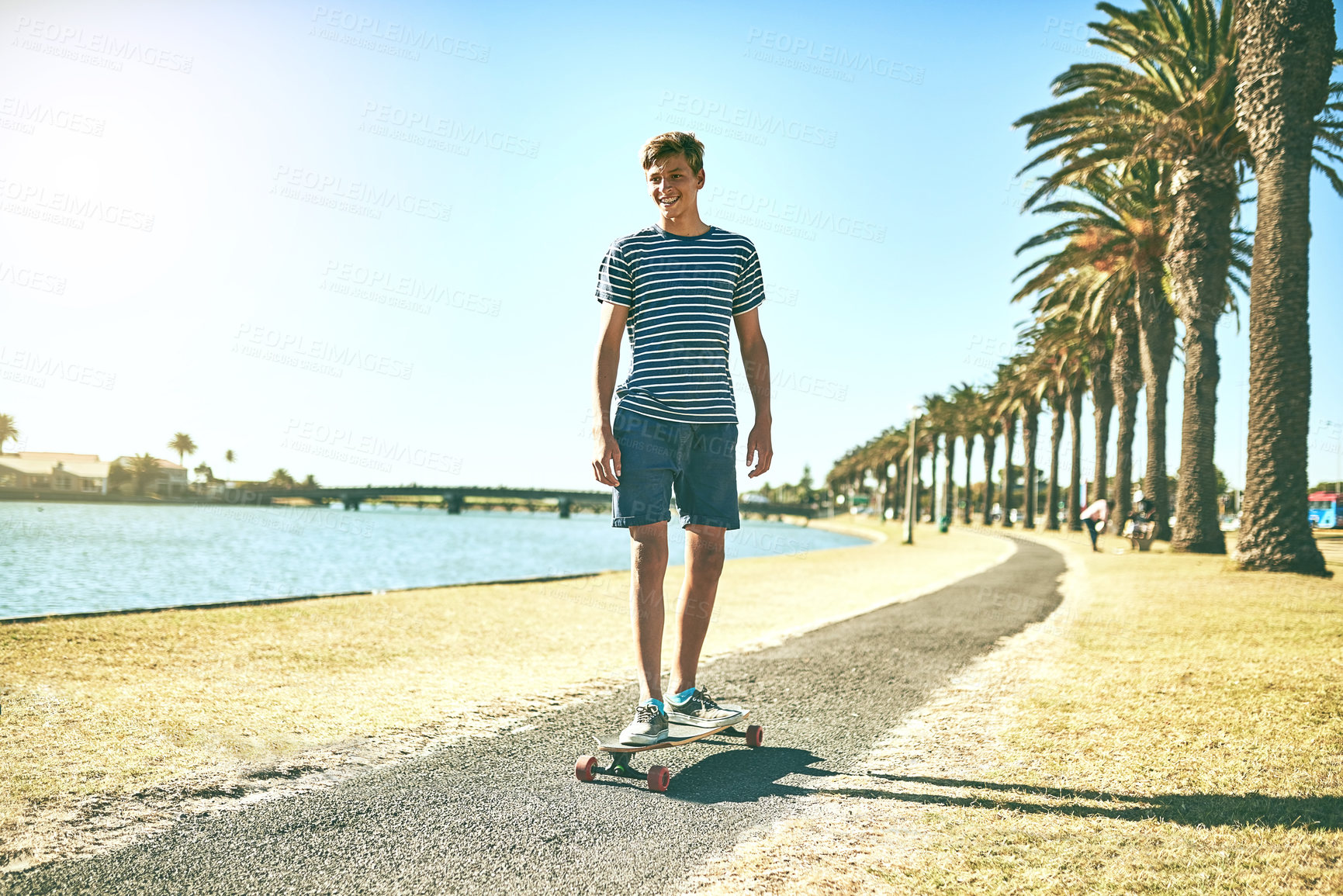 Buy stock photo Happy boy, skateboard and road with pathway in nature for cruise, travel or fun outdoor activity by lagoon. Young, male person or teenager with smile for ride, hobby or skating on street or sidewalk