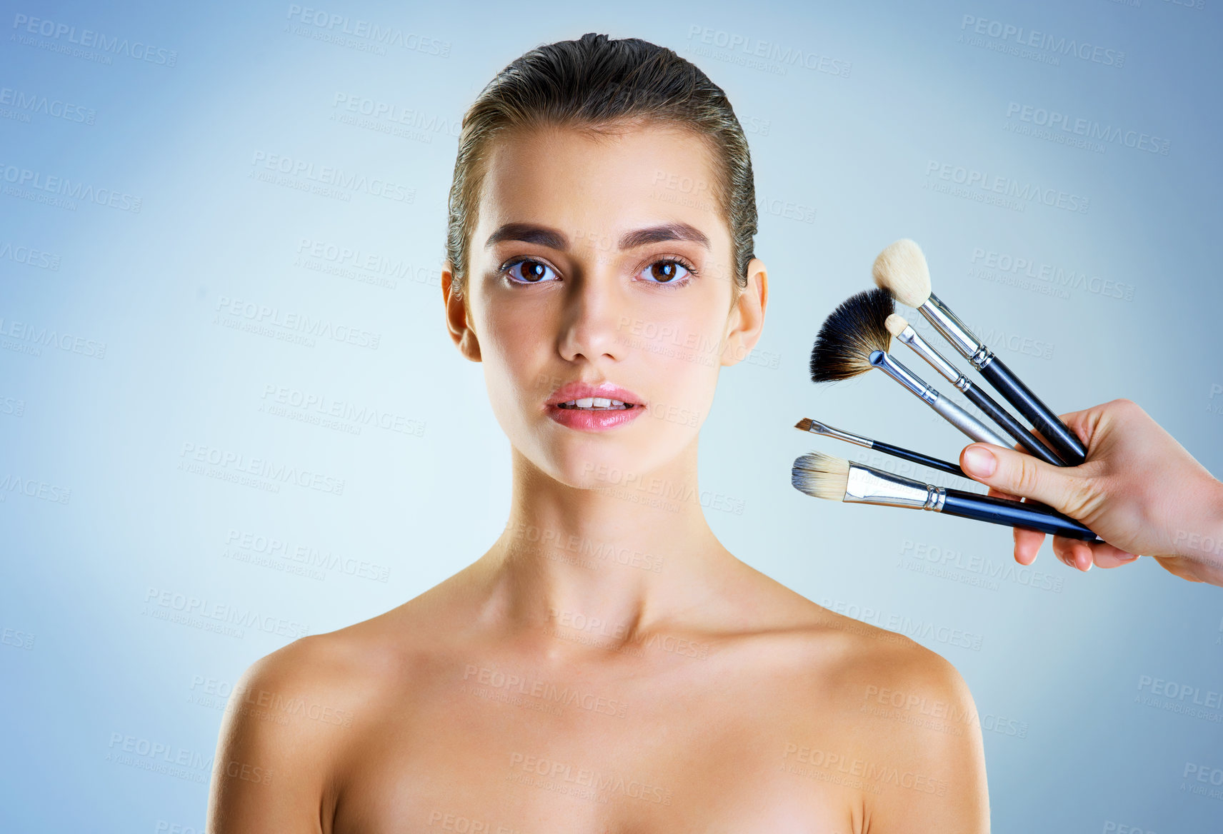 Buy stock photo Studio portrait of a hand holding makeup brushes next to a beautiful young woman against a blue background
