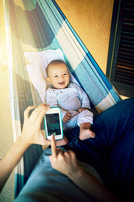 Buy stock photo Shot of an unrecognizable man taking a photo of his baby boy with a cellphone with him lying on a hammock outside at home during the day