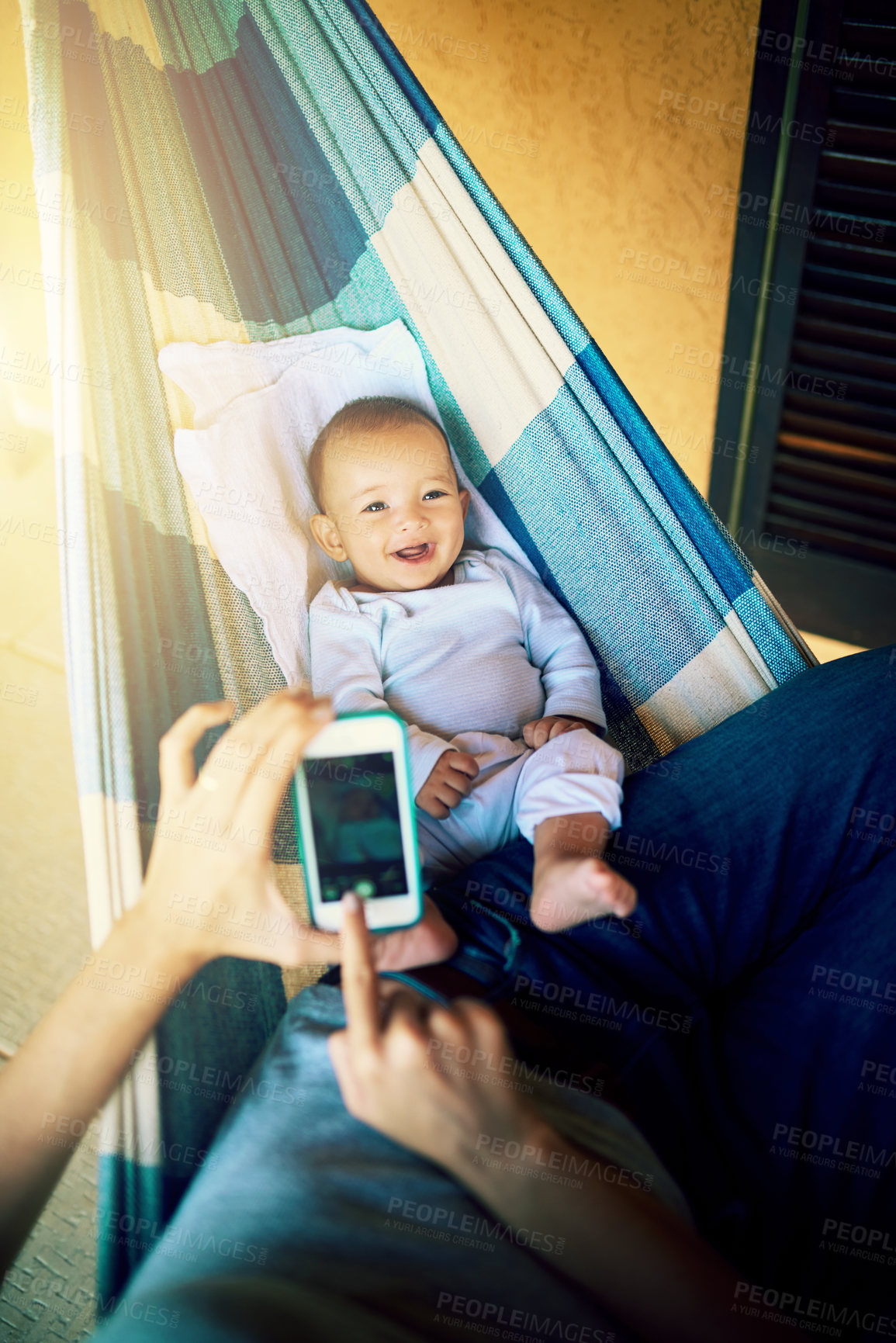 Buy stock photo Shot of an unrecognizable man taking a photo of his baby boy with a cellphone with him lying on a hammock outside at home during the day