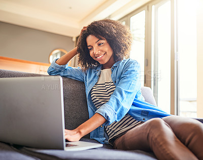 Buy stock photo Shot of a cheerful young woman working on a laptop while being seated on a couch at home during the day