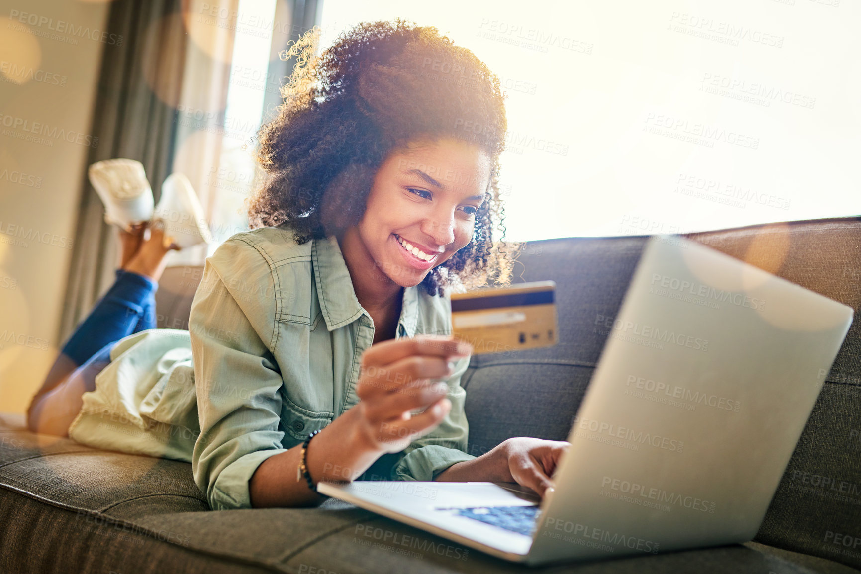 Buy stock photo Shot of a cheerful young woman doing online shopping while lying on a couch at home during the day