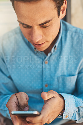 Buy stock photo Shot of a focused young man texting on his cellphone while being seated outside during the day