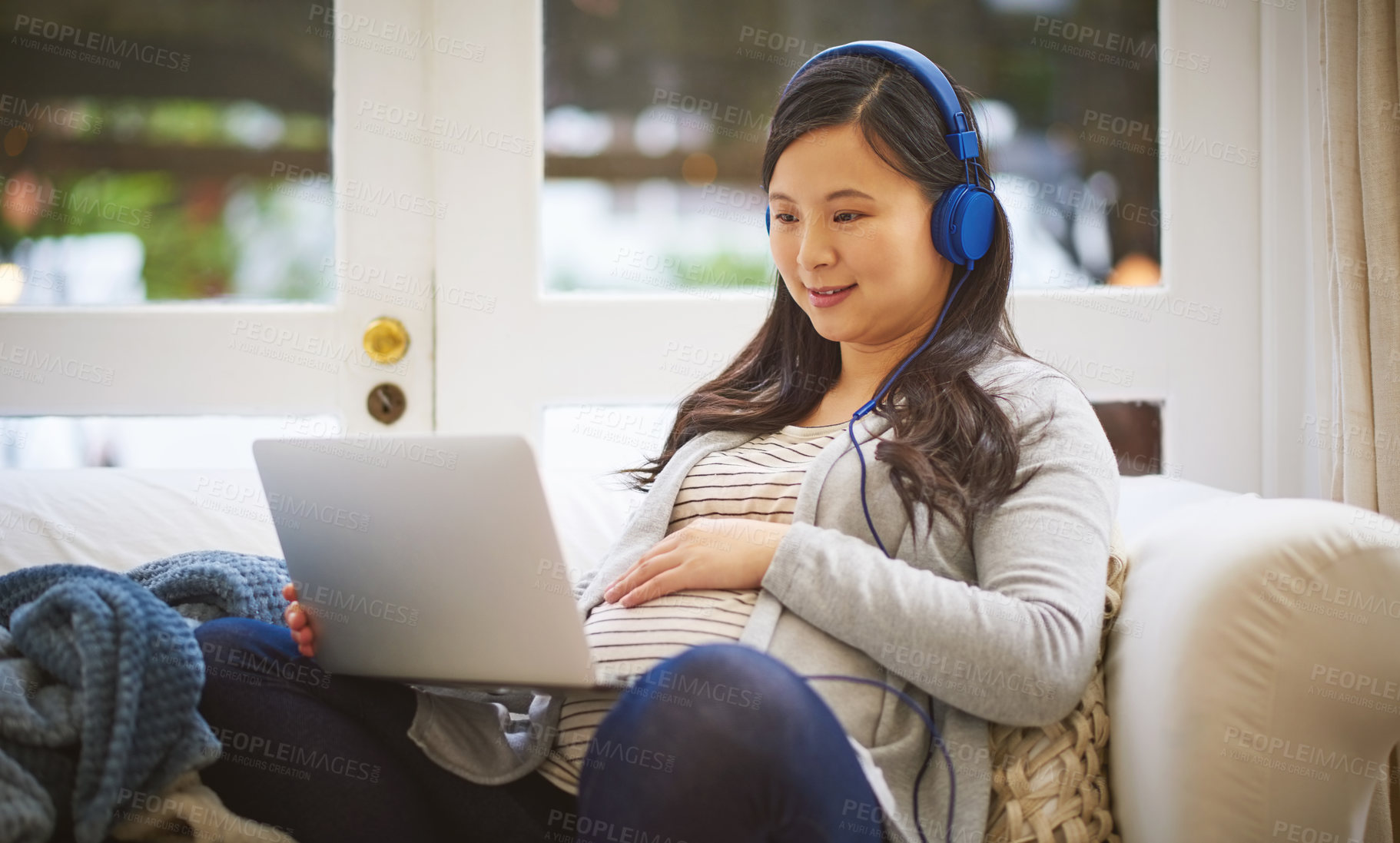 Buy stock photo Shot of a pregnant woman wearing headphones while using a laptop at home