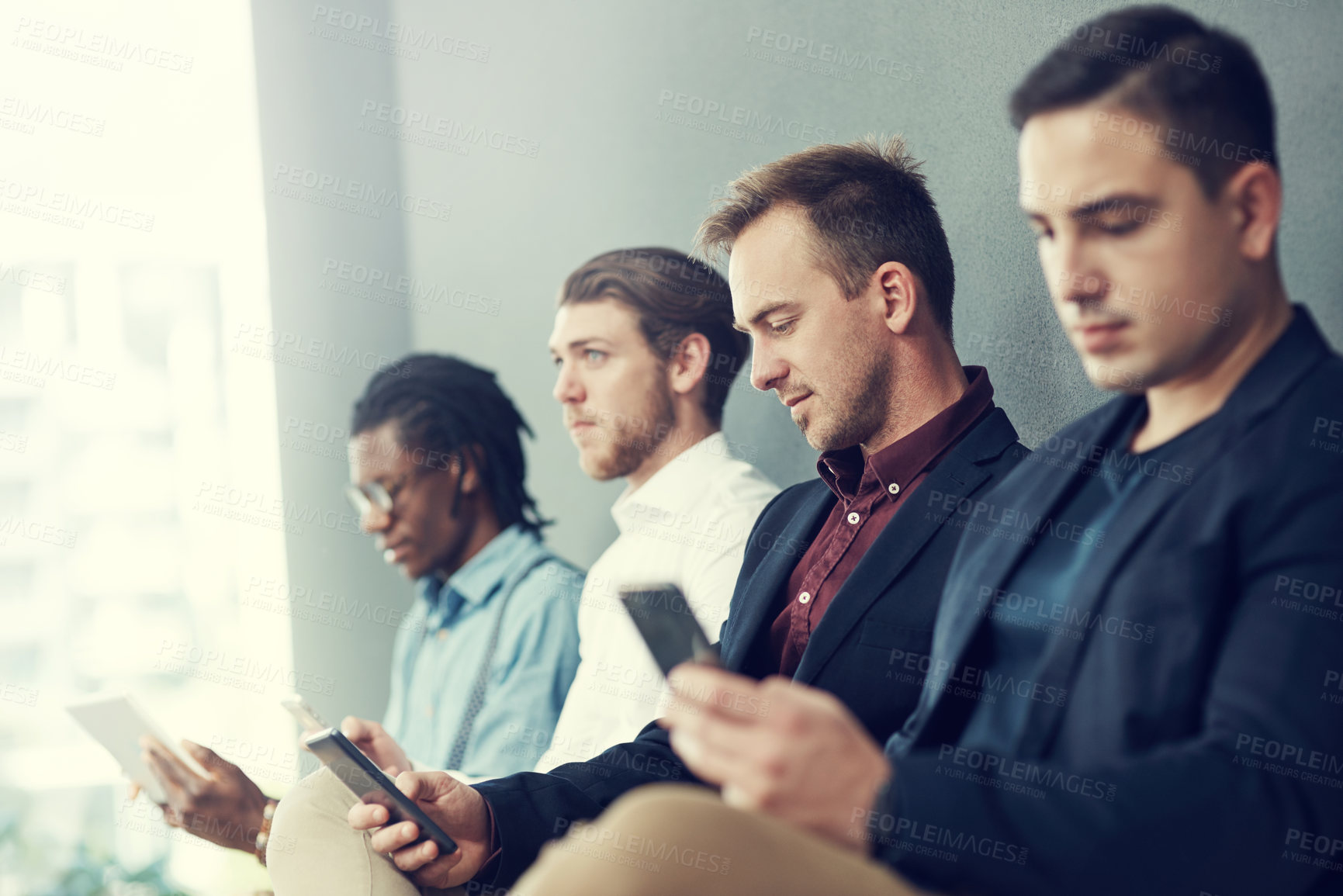 Buy stock photo Shot of a group of businessmen using different wireless devices while waiting in line for an interview