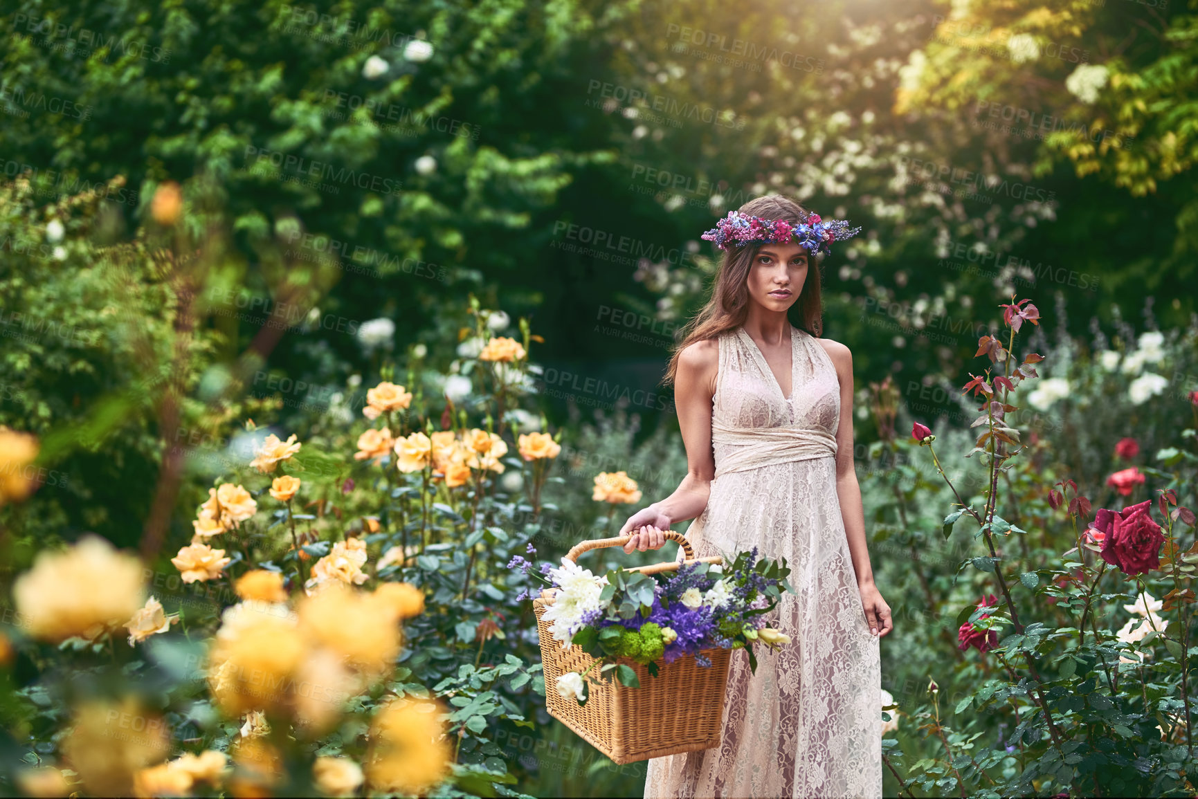 Buy stock photo Shot of a beautiful young woman wearing a floral head wreath and holding a basket full of flowers in nature