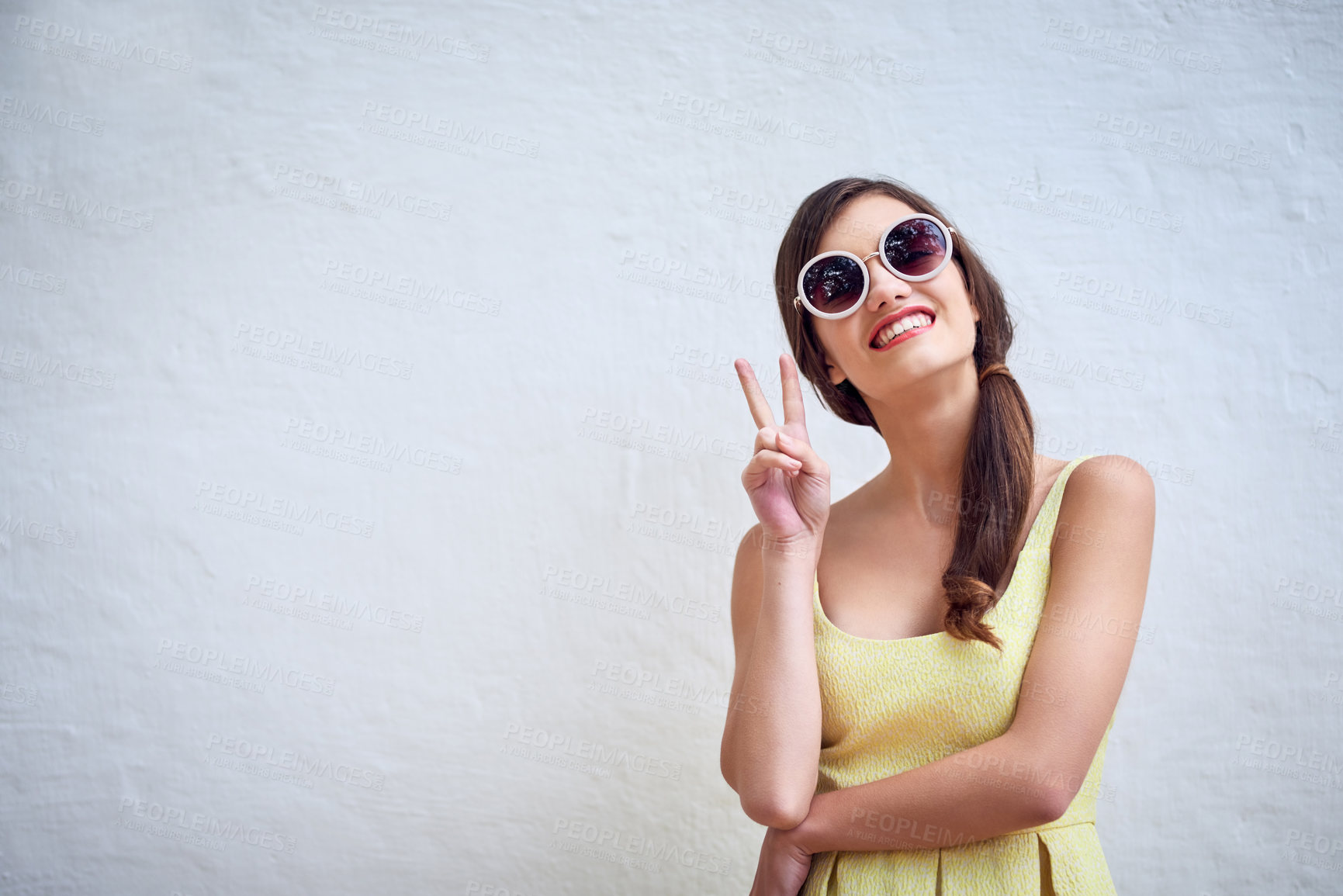 Buy stock photo Studio portrait of a cheerful young woman wearing sunglasses while showing a peace sign against a grey background