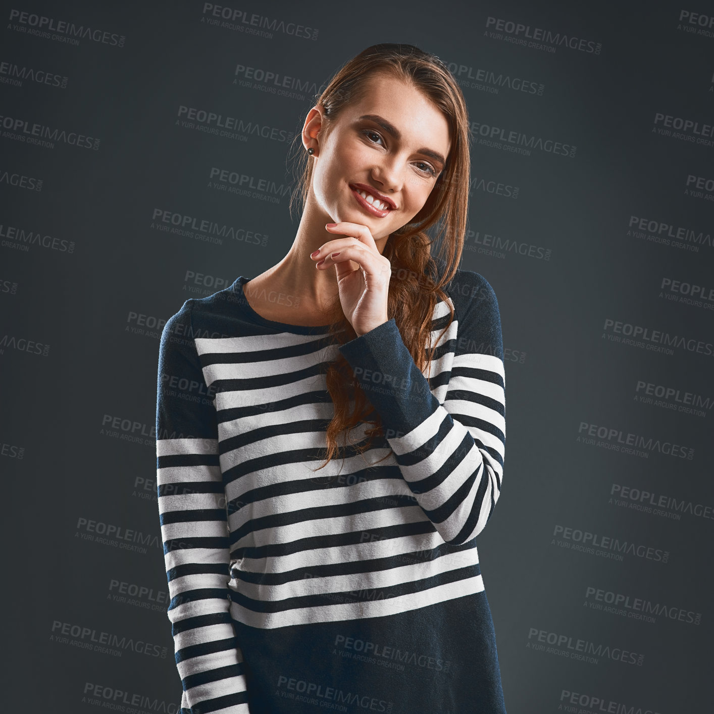 Buy stock photo Studio portrait of a cheerful young woman holding her chin while standing against a dark background