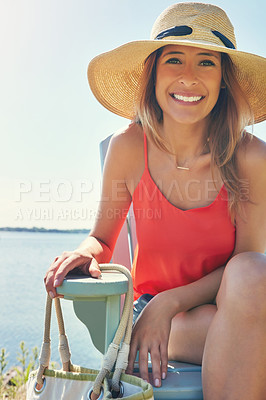 Buy stock photo Portrait of a cheerful young woman wearing a hat while being seated on a chair next to a lake outside in the sun