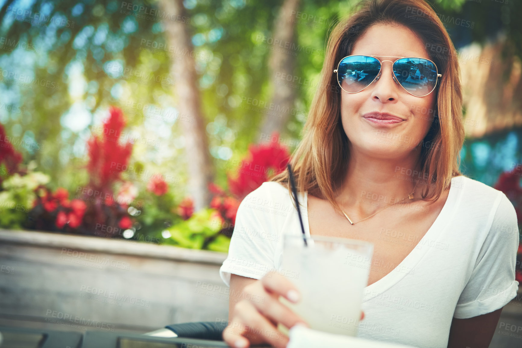 Buy stock photo Portrait of a cheerful young woman wearing sunglasses and enjoying a cold beverage while being seated at a restaurant outside during the day