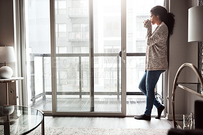 Buy stock photo Shot of a cheerful young woman drinking coffee while looking through a window inside at home during the day