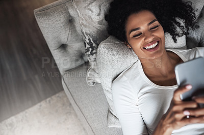 Buy stock photo Shot of a cheerful young woman relaxing on a couch while texting on her cellphone at home during the day