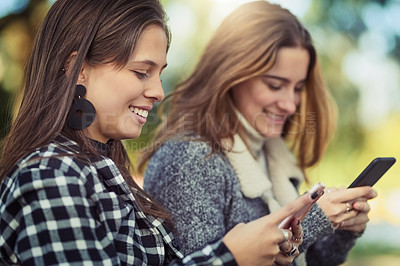 Buy stock photo Cropped shot of two attractive young women using their cellphones while sitting in the park