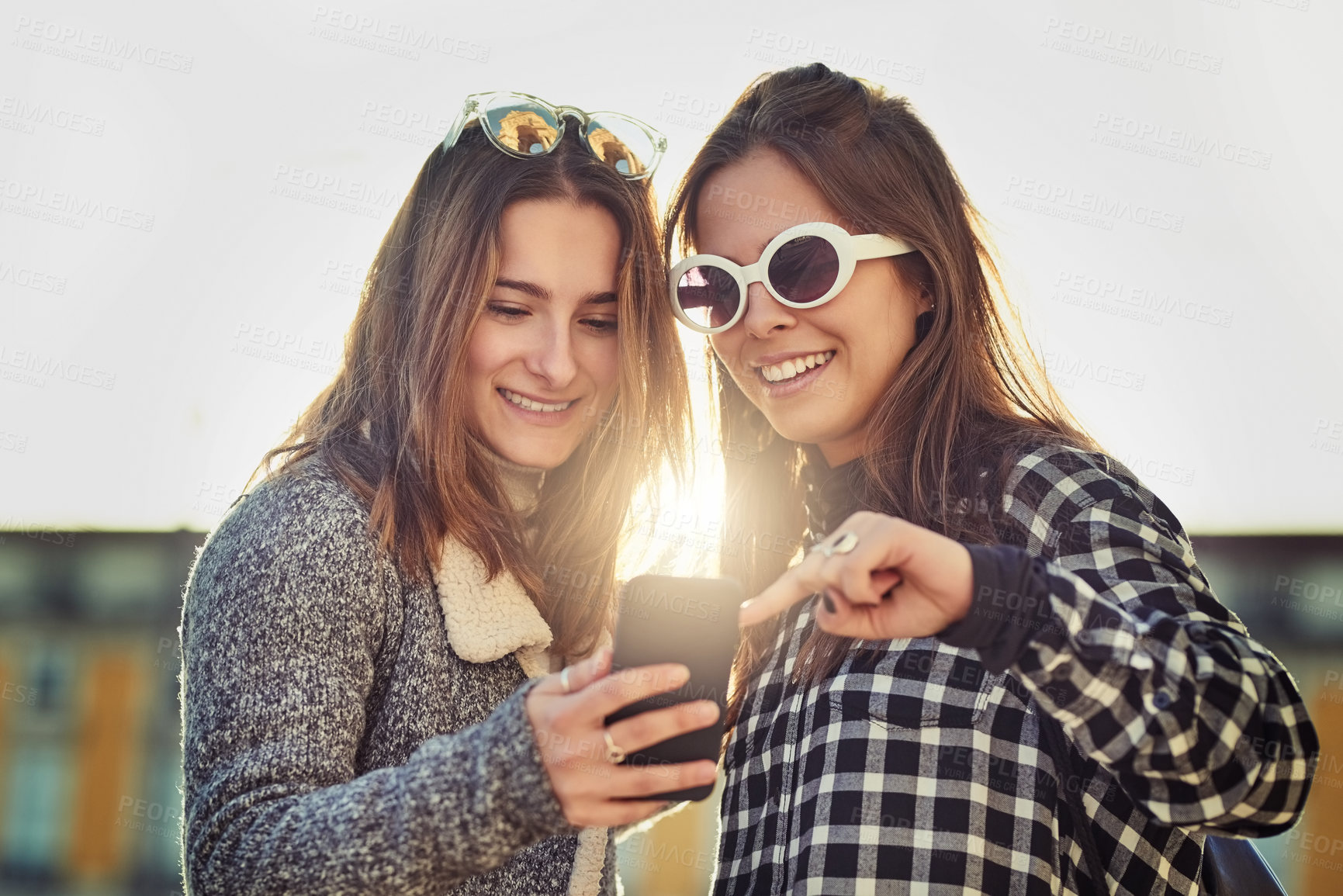Buy stock photo Cropped shot of two attractive young women using a cellphone while out in the city