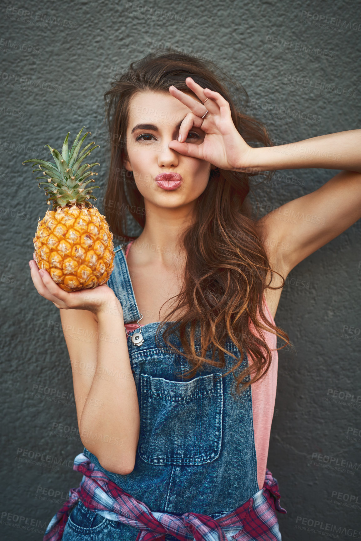 Buy stock photo Portrait of a carefree young woman showing a hand gesture on her face while holding a pineapple against a grey background