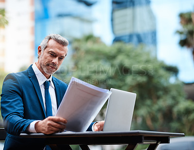 Buy stock photo Shot of a handsome mature businessman in corporate attire going over a document and using a laptop outside during the day