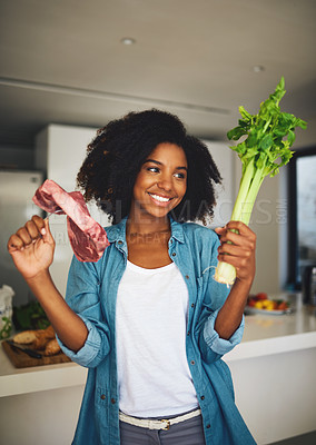 Buy stock photo Shot of a cheerful young woman holding celery in one hand and a piece of meat in the other while standing in the kitchen at home