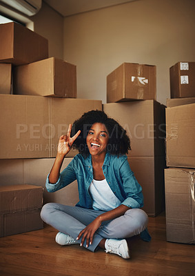 Buy stock photo Portrait of a cheerful young woman seated on the floor while being surrounded by boxes and showing the peace sign in side at home