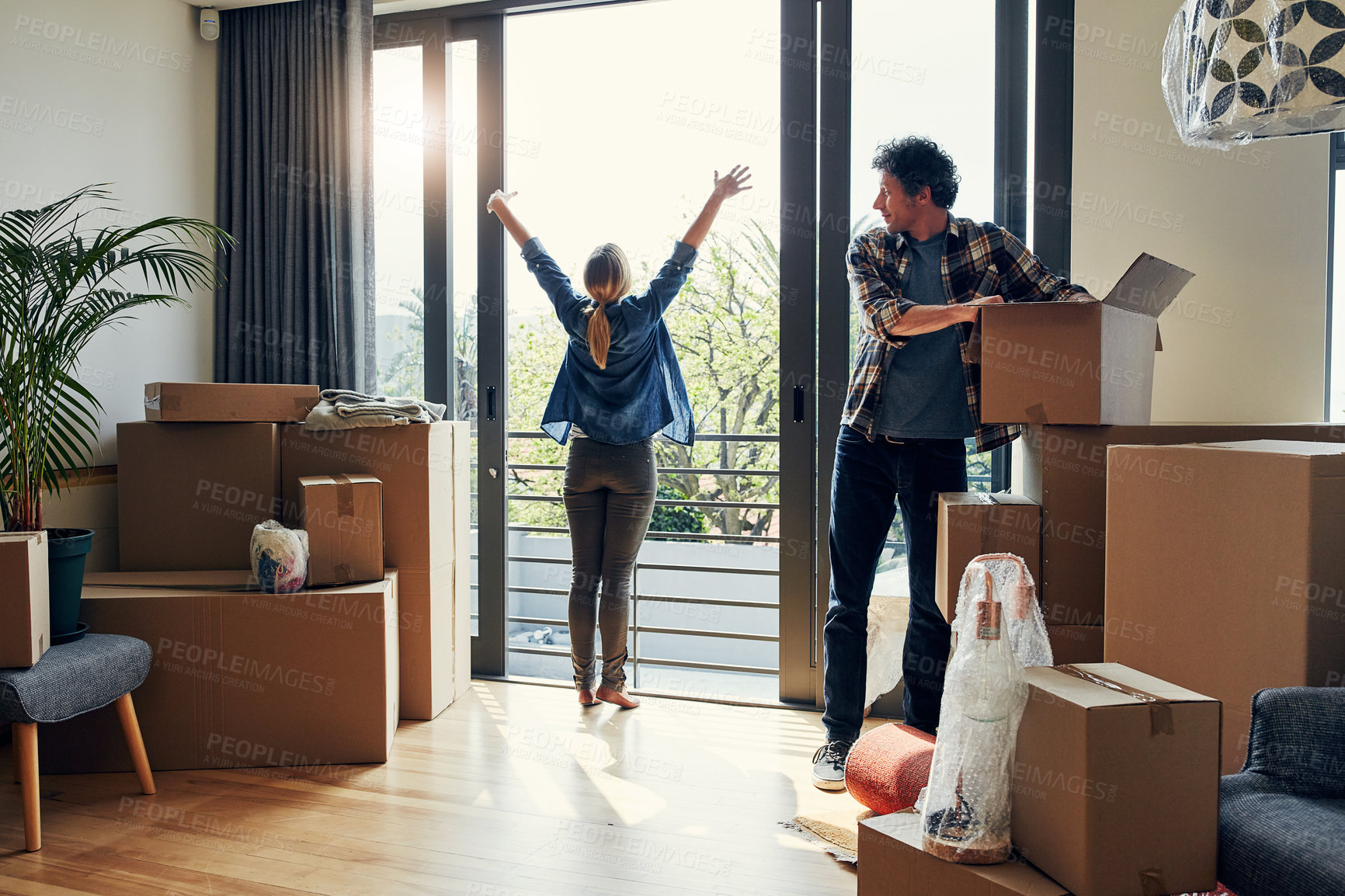 Buy stock photo Shot of a cheerful middle aged woman stretching her arms out on her balcony while her husband looks at her and packs out boxes on moving day
