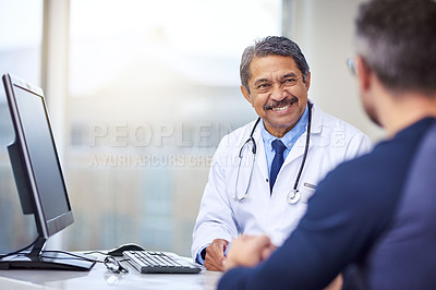 Buy stock photo Shot of a cheerful mature male doctor seated at his desk while consulting a patient inside a hospital during the day