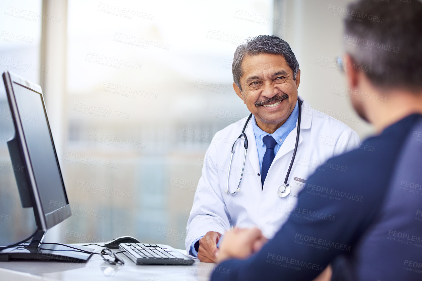 Buy stock photo Shot of a cheerful mature male doctor seated at his desk while consulting a patient inside a hospital during the day