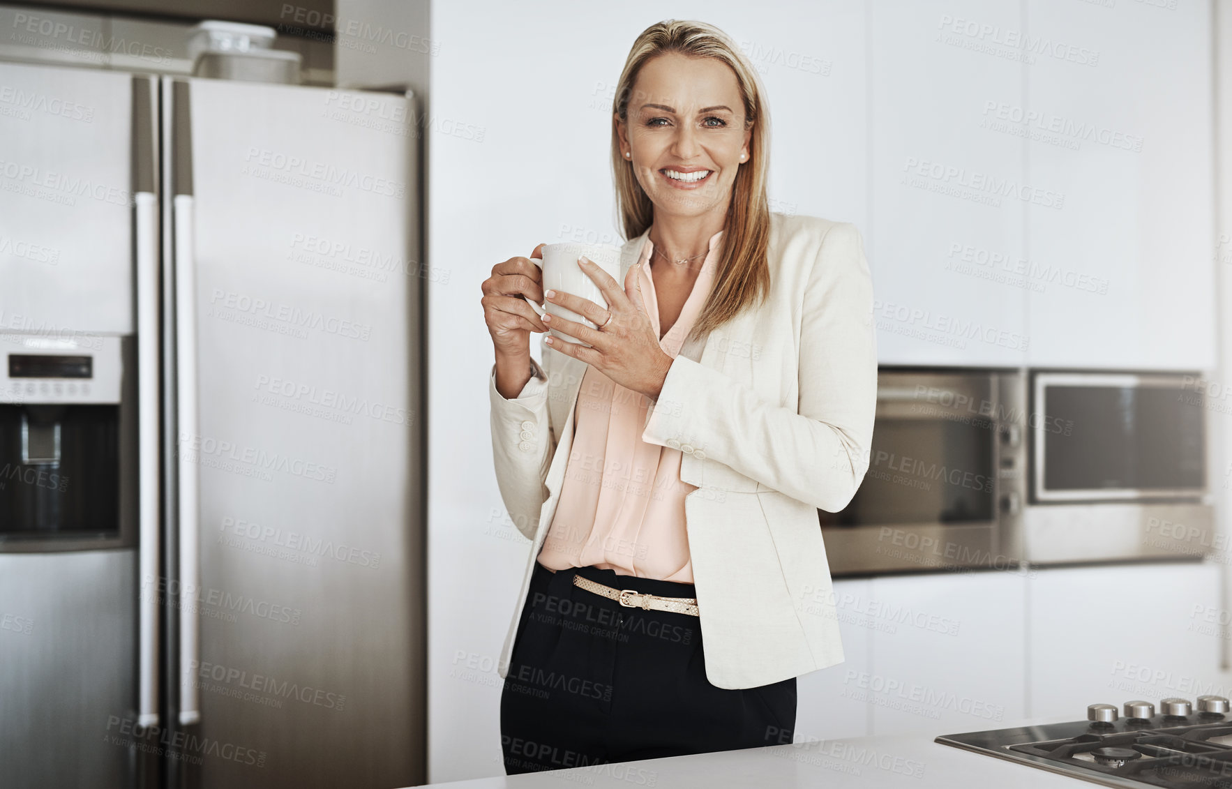 Buy stock photo Portrait of a cheerful middle aged businesswoman standing behind a counter while drinking a cup of coffee inside of a kitchen during the day