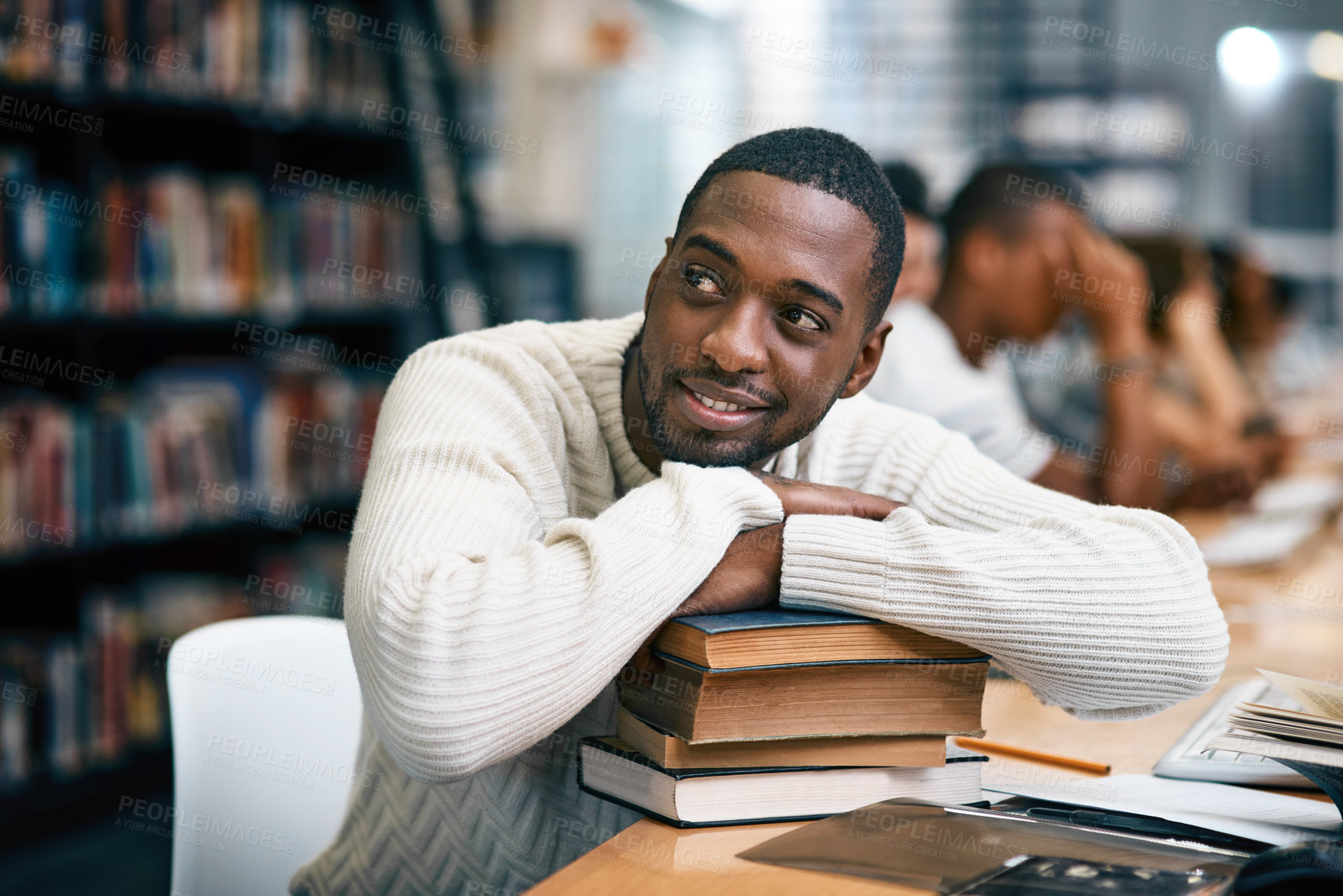Buy stock photo Shot of a young man resting on a pile of books in a college library and looking thoughtful