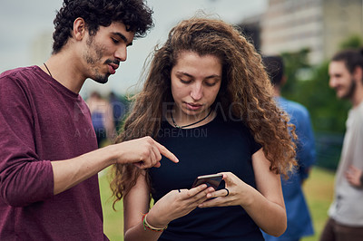Buy stock photo Shot of two cheerful young friends browsing on a cellphone together while standing in a park outside during the day