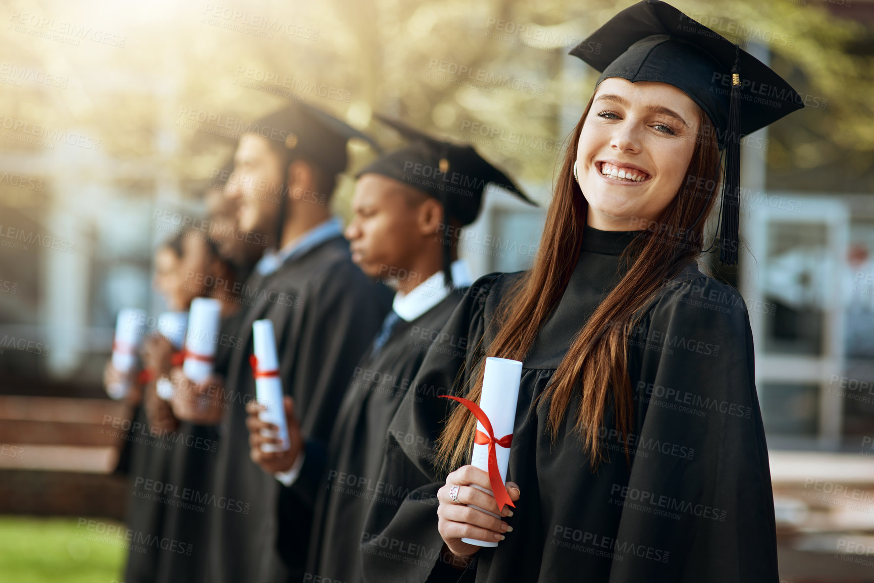 Buy stock photo Portrait of a young woman holding her diploma while standing with her fellow students on graduation day
