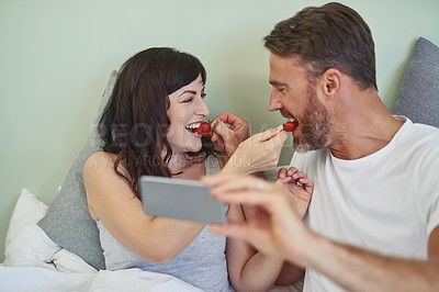 Buy stock photo Shot of a cheerful young couple sitting in bed while enjoying breakfast together and taking a selfie during morning hours