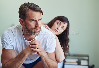 Buy stock photo Shot of a unhappy looking middle aged man looking into the distance while his partner tries to comfort him in the bedroom