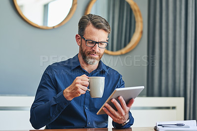 Buy stock photo Shot of a focused middle aged businessman browsing on a digital tablet while enjoying a cup of coffee at home during the day