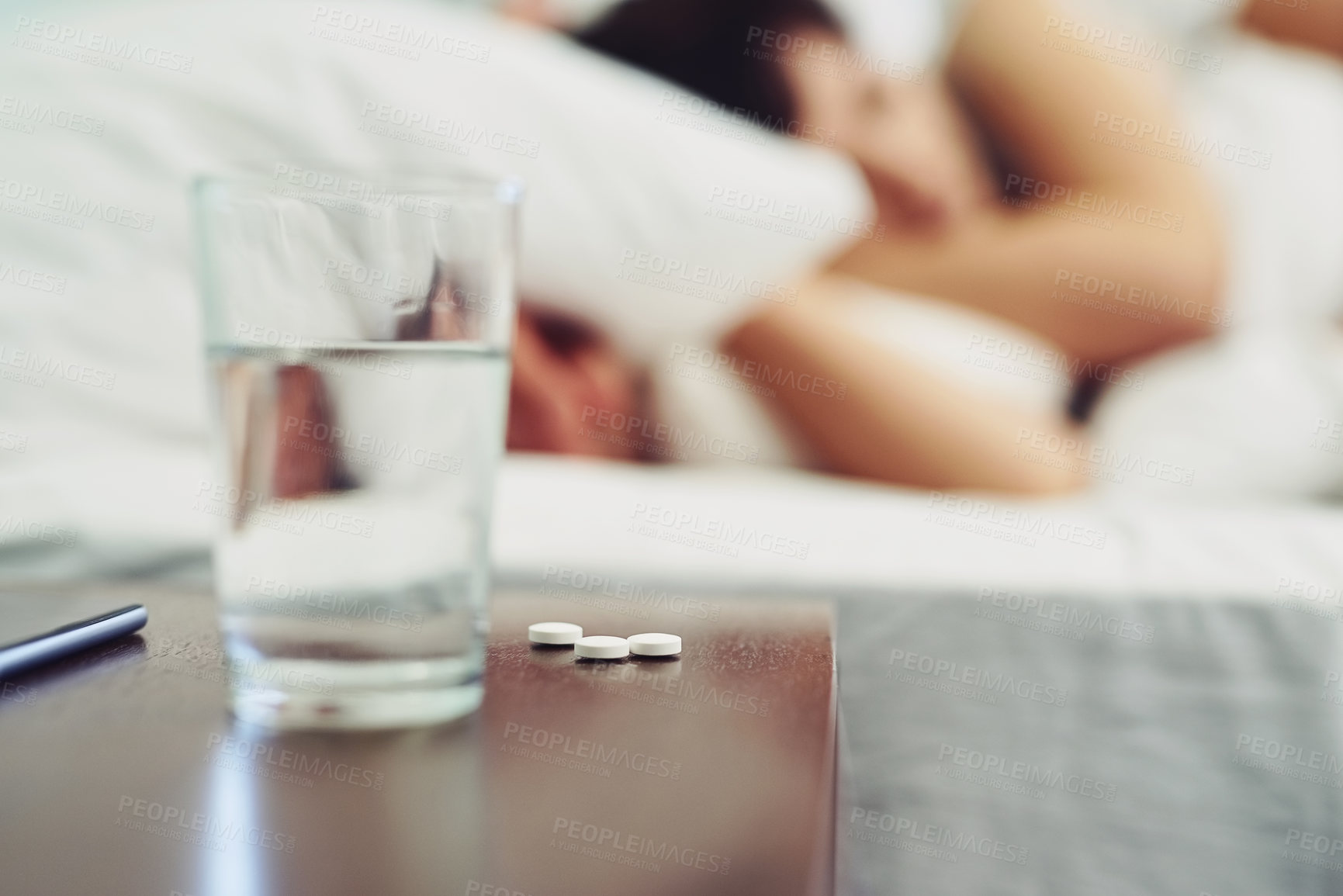 Buy stock photo Shot of a glass of water standing next to a couple of pills on a bedside table at home during the day