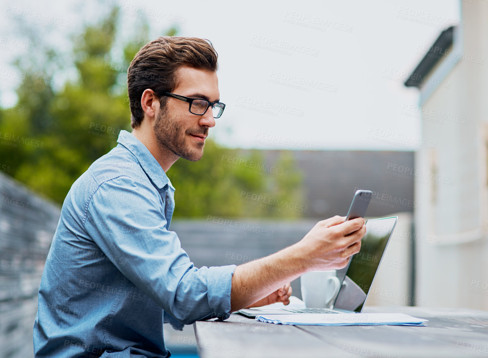 Buy stock photo Shot of a handsome young man using a laptop and cellphone outdoors