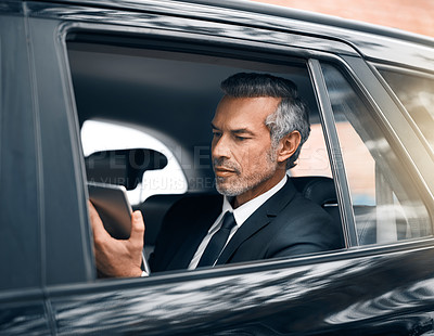 Buy stock photo Cropped shot of a handsome mature businessman using a tablet while sitting in the backseat of a car during his morning commute