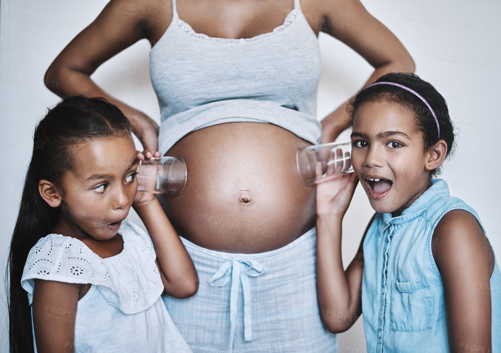 Buy stock photo Portrait of two cheerful little girls standing next their mother while each putting a glass on her pregnant belly to listen for movement at home