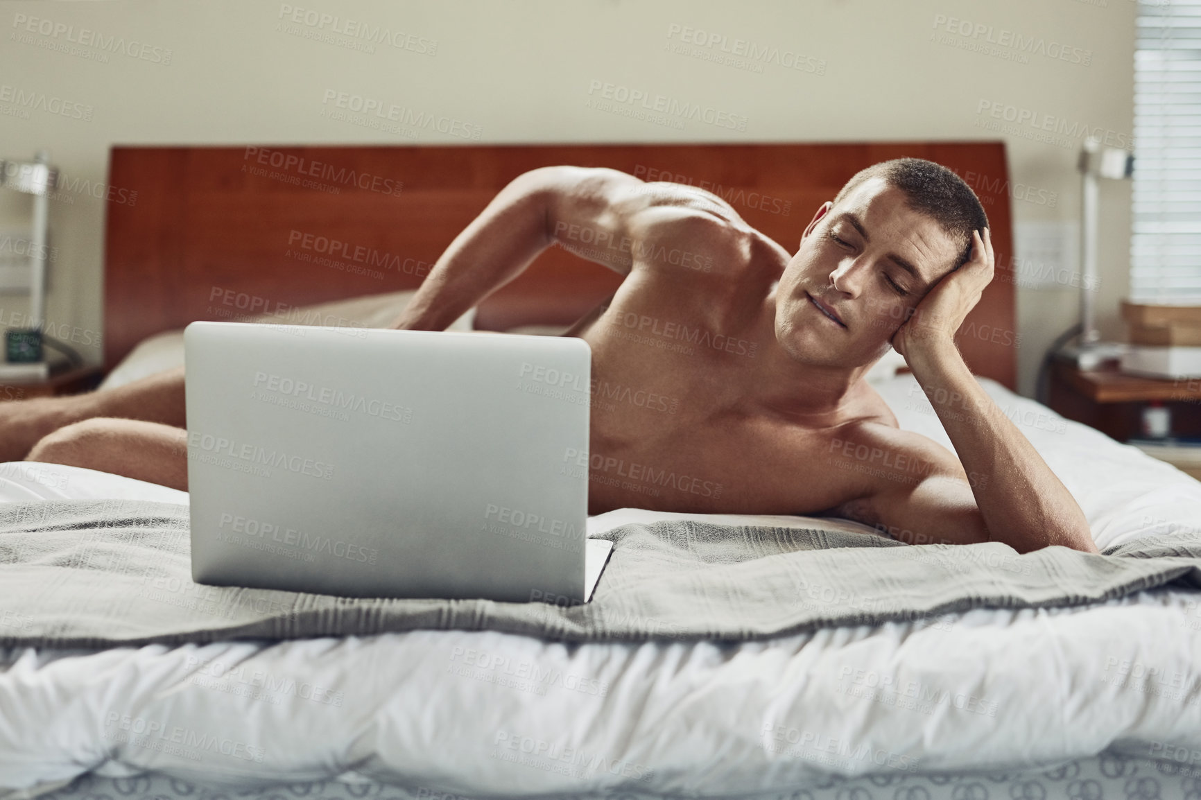 Buy stock photo Shot of a carefree young naked man lying on his bed while being busy with his laptop at home during the day