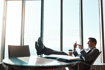 Buy stock photo Shot of a young businessman using a cellphone while relaxing with his feet up on a table in an office