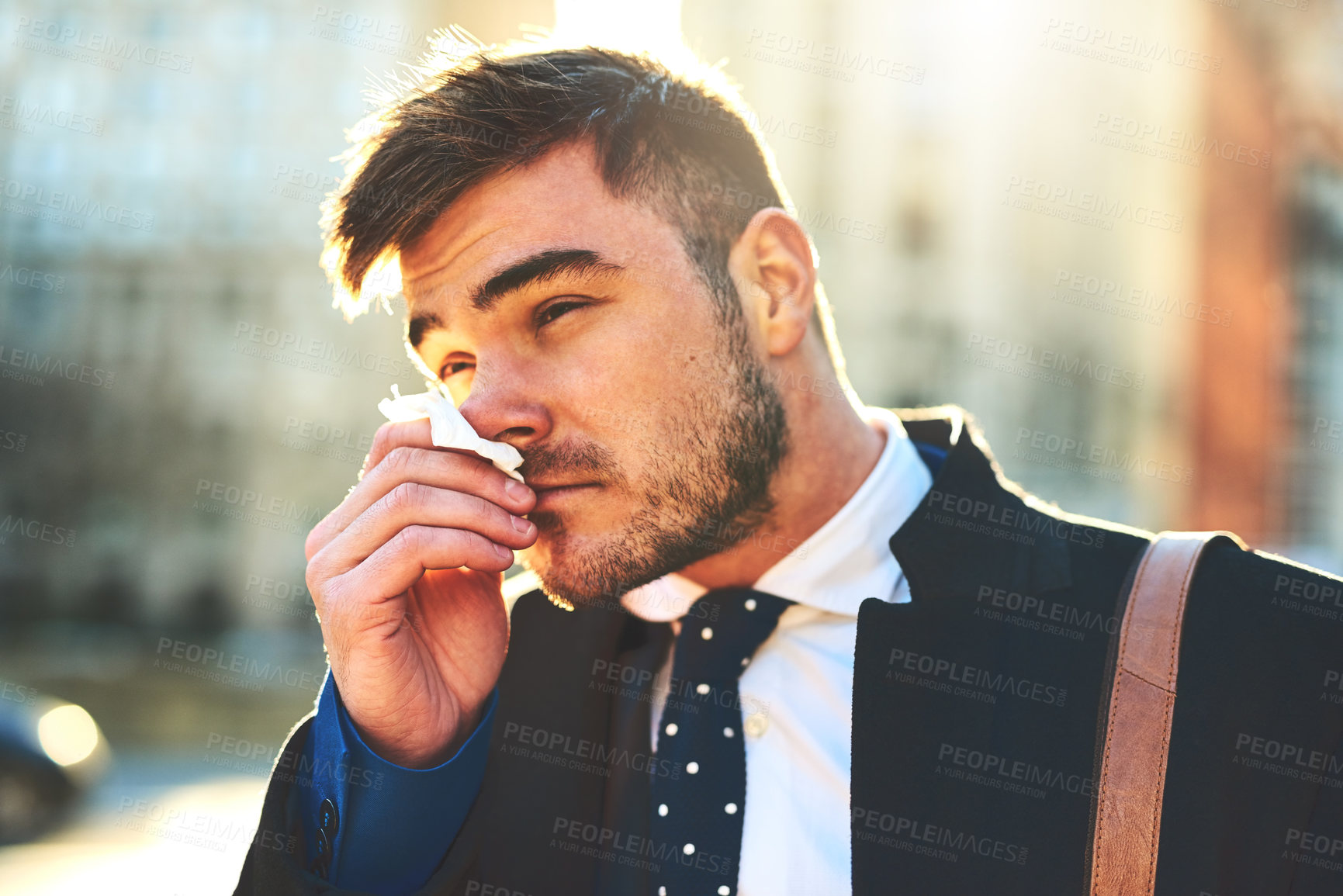 Buy stock photo Shot of a irritated looking young man blowing his nose with a tissue while walking the busy streets of the city in the morning