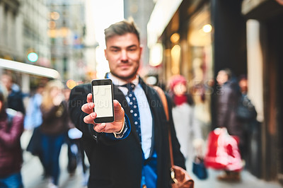 Buy stock photo Portrait of a cheerful young man holding up a cellphone and showing the screen to the camera outside in the city during the day