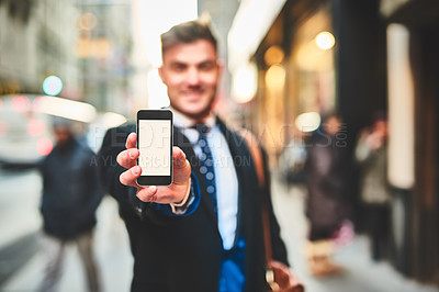 Buy stock photo Portrait of a cheerful young man holding up a cellphone and showing the screen to the camera outside in the city during the day