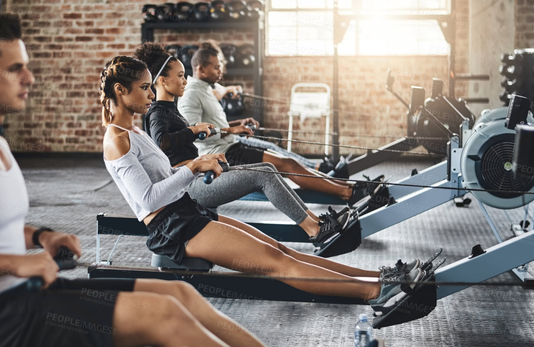 Buy stock photo Shot of a group of young people working out with rowing machines in the gym
